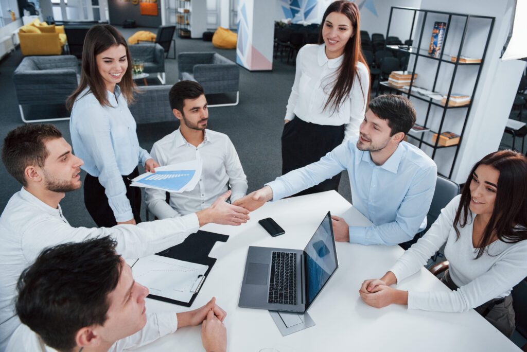 Handshake for success deal. Top view of office workers in classic wear sitting near the table using laptop and documents Know Us